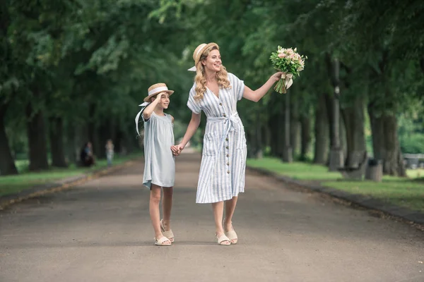 母亲和女儿在稻草帽子与花束牵手和步行在绿色公园的路径 — 图库照片