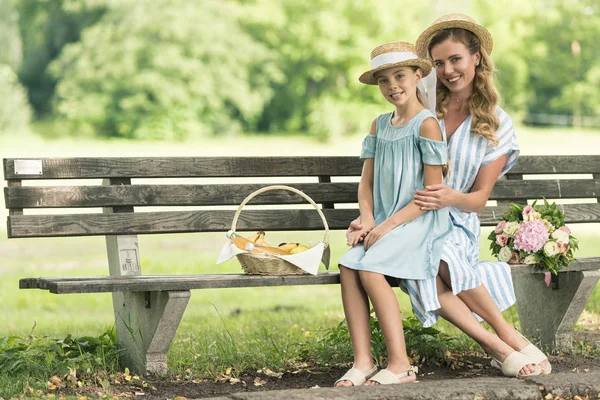 Stock image attractive mother and daughter in straw hats with wicker basket sitting on bench