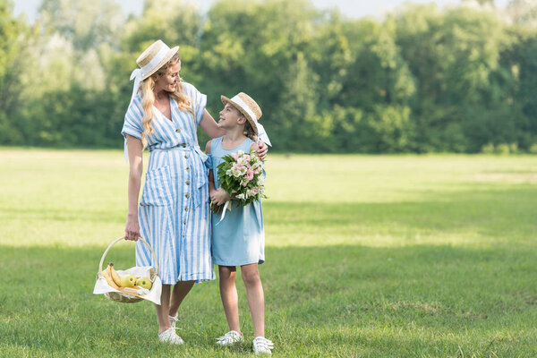 beautiful mother and daughter with wicker basket and bouquet walking on green lawn