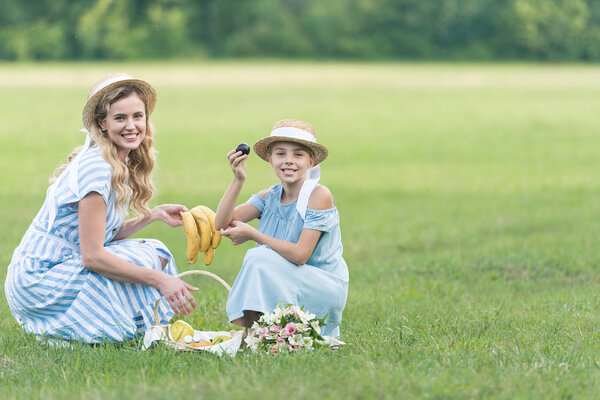 smiling mom and daughter having picnic with fruits and bouquet on green lawn