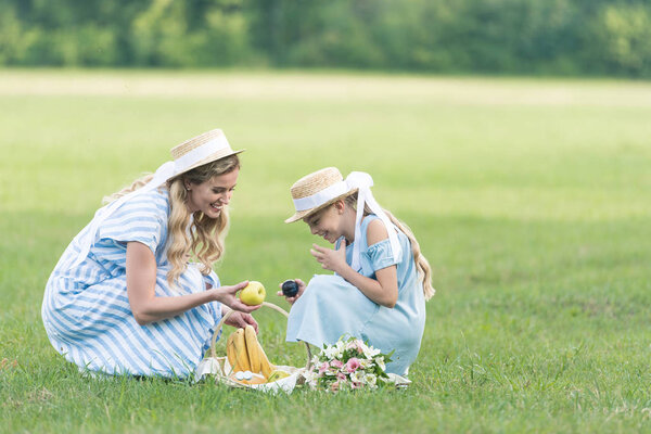 beautiful mother and happy daughter having picnic with fruits and bouquet on green lawn