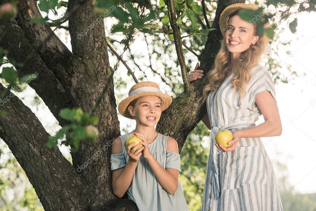 happy mother and daughter in straw hats holding appples and standing near tree