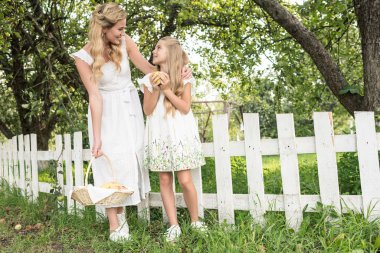 blonde mother and daughter with fruits in wicker basket posing near white fence in garden clipart