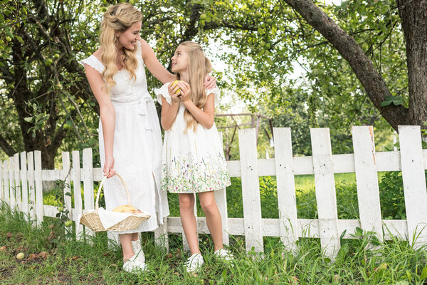 blonde mother and daughter with fruits in wicker basket posing near white fence in garden