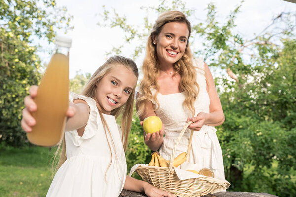 Улыбающаяся дочь держит бутылку свежего сока и красивую мать держа плетеную корзину с фруктами в саду
