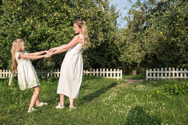 attractive mother and daughter in white dresses holding hands and twisting in orchard
