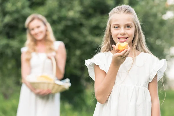 Adorable Daughter Eating Peach While Mother Holding Wicker Basket Background — Stock Photo, Image