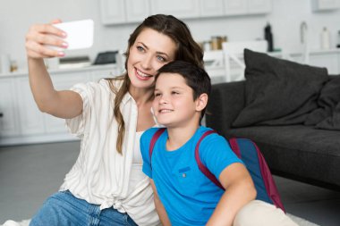 Anne ve oğlu selfie Smartphone cep telefonu ile birlikte evde ilk okul gününde almak sırt çantası ile gülümseyen
