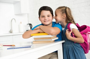 portrait of smiling boy at table with books and little sister with backpack telling secret near by at home clipart