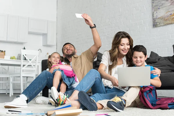 微笑的父亲和女儿采取自拍智能手机 而母亲和儿子使用笔记本电脑一起在家里 — 图库照片