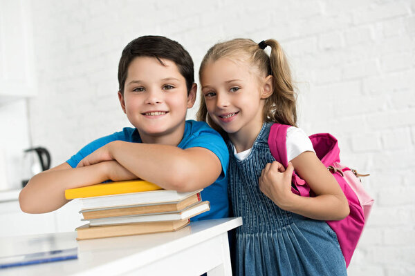 portrait of smiling boy at table with books and little sister with backpack near by at home