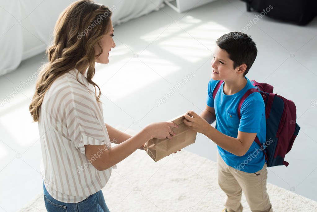 side view of mother giving paper package with meal to son with backpack at home, back to school concept