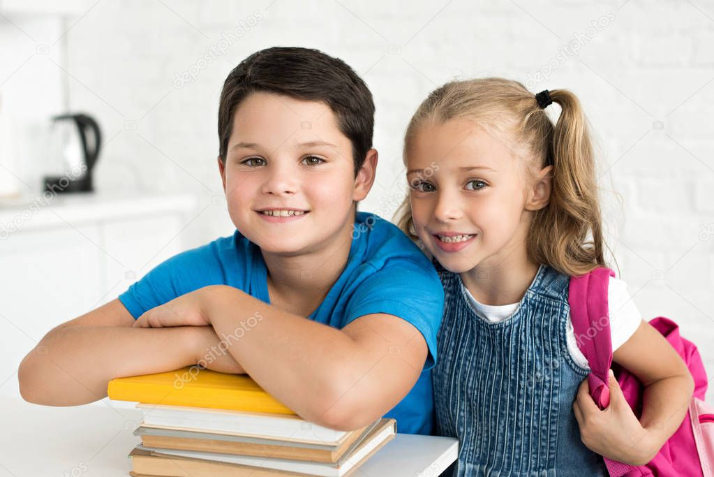 portrait of smiling boy at table with books and little sister with backpack near by at home