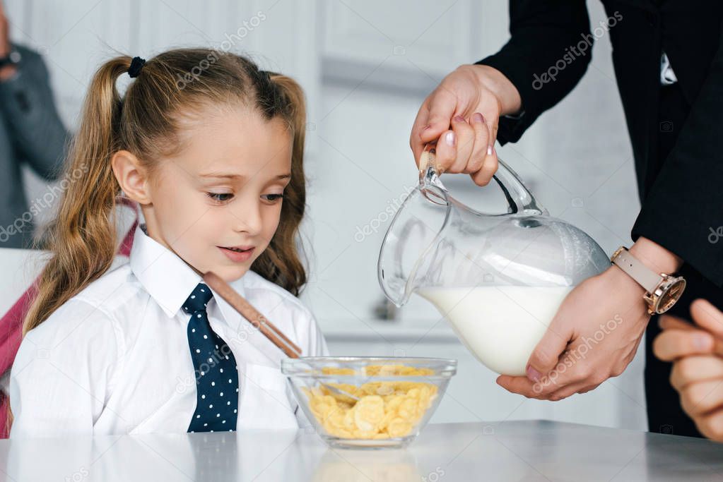 selective focus of kid in school uniform at table with breakfast and mother pouring milk into bowl with crunches at home, back to school concept