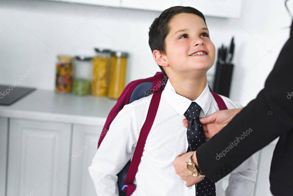 partial view of mother in suit tying sons tie in kitchen at home, back to school concept