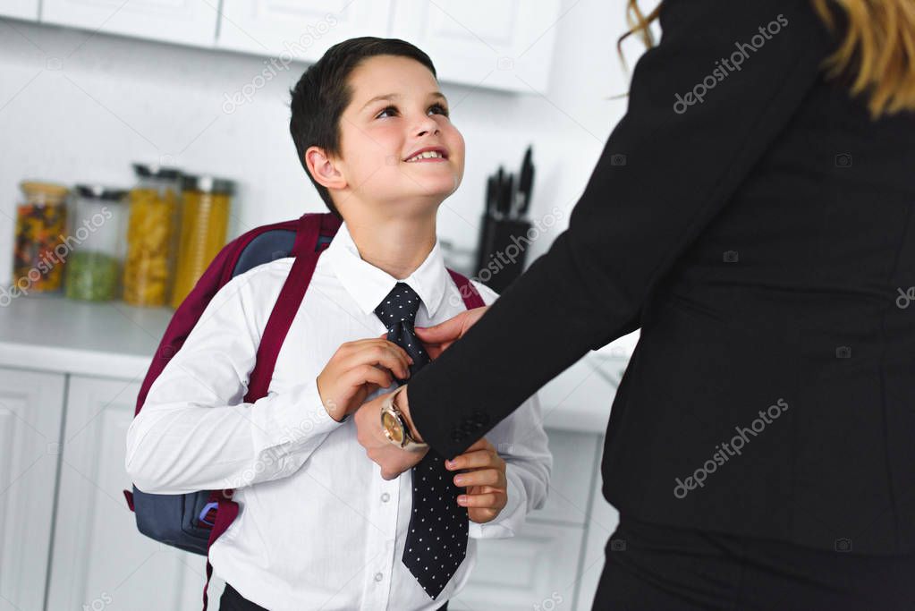 partial view of mother in suit and son in school uniform with backpack in kitchen at home, back to school concept