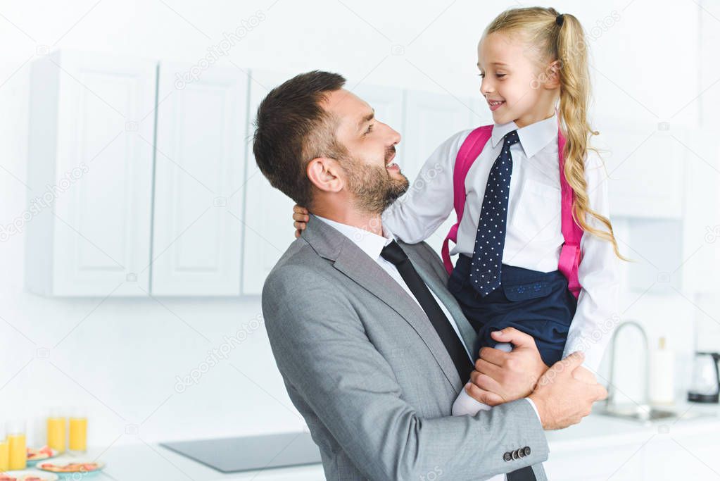 portrait of happy father in suit holding daughter in school uniform with backpack on hands in kitchen, back to school concept