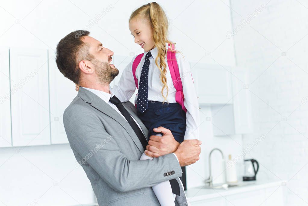 portrait of happy father in suit holding daughter in school uniform with backpack on hands in kitchen, back to school concept