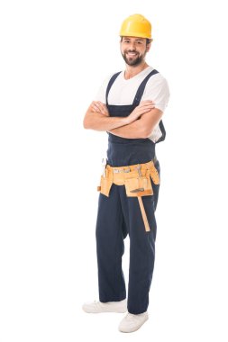 full length view of handsome happy workman in hard hat and tool belt standing with crossed arms and smiling at camera isolated on white clipart
