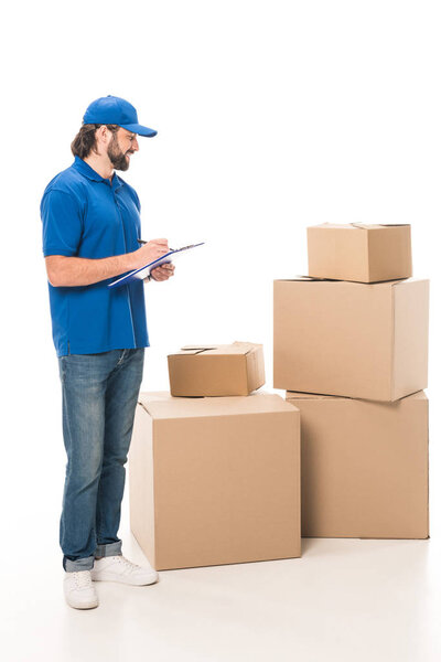 full length view of smiling delivery man writing on clipboard and looking at cardboard boxes isolated on white