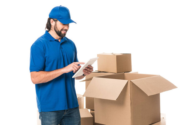 smiling delivery man using digital tablet while standing near boxes isolated on white
