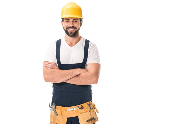 handsome happy workman in hard hat and tool belt standing with crossed arms and smiling at camera isolated on white