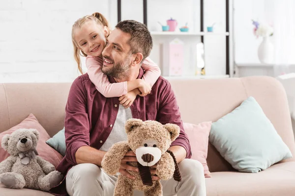 女儿抱着爸爸在沙发上与泰迪熊在家里 — 图库照片