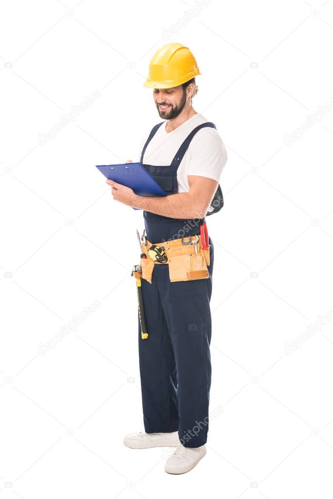 full length view of smiling workman in tool belt writing on clipboard isolated on white