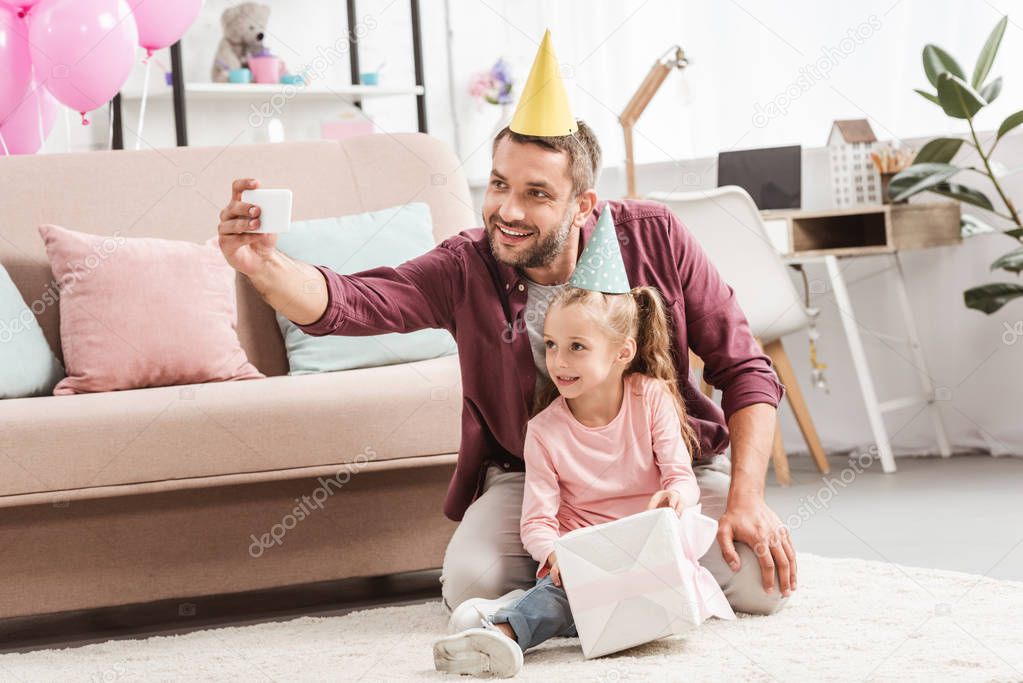 smiling father and daughter in party hats taking selfie with gift for birthday