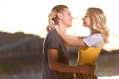 side view of smiling couple hugging and looking at each other on river beach in evening clipart