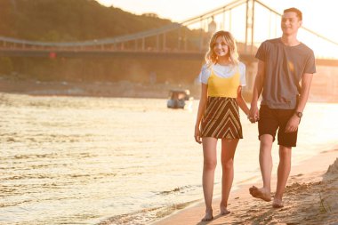 Elele ve çıplak ayakla akşam nehir kumsalda yürüyen genç çift gülümseyen