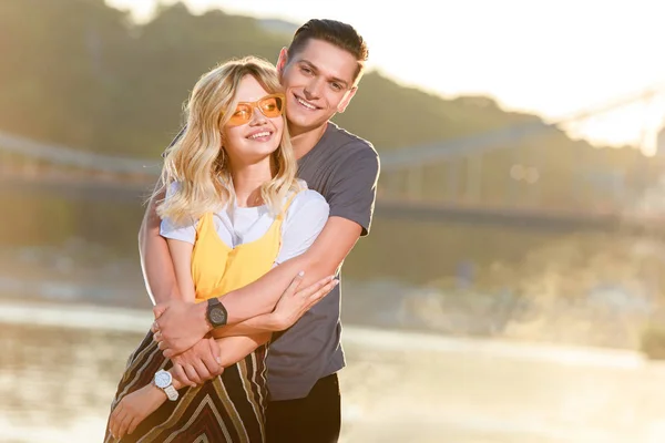 Smiling Boyfriend Hugging Girlfriend River Beach Evening Looking Camera — Free Stock Photo