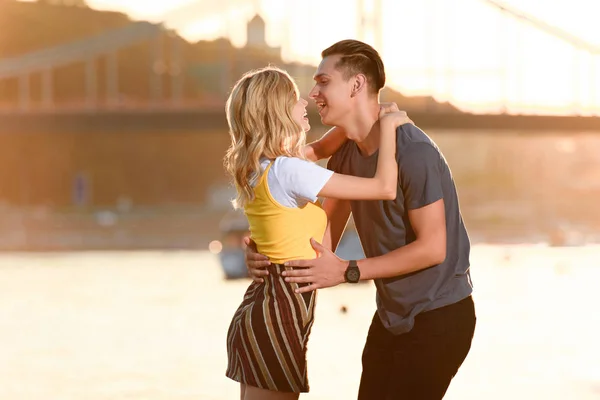 笑的年轻夫妇的侧面观看晚上在河边海滩亲吻 — 免费的图库照片