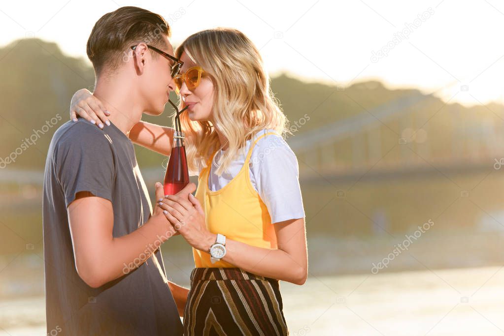 couple hugging and drinking beverage with two plastic straws from one bottle on river beach in evening