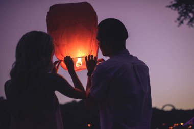 back view of young couple launching sky lantern on river beach in evening clipart