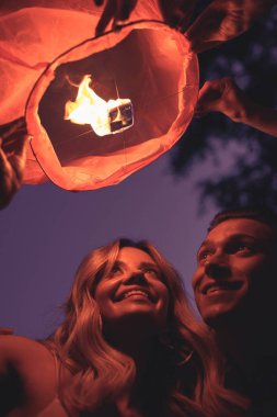 low angle view of smiling young couple launching sky lantern on river beach in evening clipart