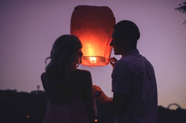 silhouettes of couple launching sky lantern on river beach in evening and looking at each other clipart