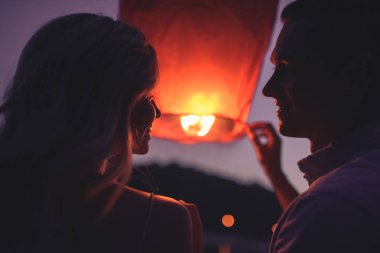 portrait of couple launching sky lantern on river beach in evening and looking at each other clipart