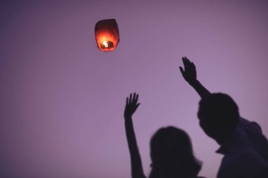 silhouettes of couple waving hands to flying sky lantern in evening clipart