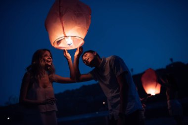 young couple looking at sky lantern on river beach in evening clipart