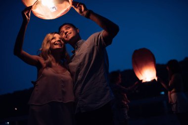 young couple launching sky lantern on river beach in evening clipart