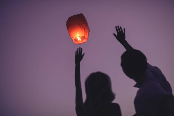 silhouettes of couple waving hands to flying chinese lantern in violet evening sky