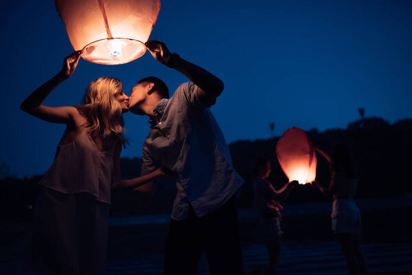 young couple kissing and launching sky lantern on river beach in evening