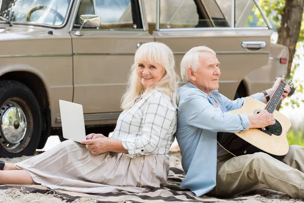 Seniorin Benutzt Laptop Während Mann Gitarre Gegen Oldtimer Spielt — kostenloses Stockfoto