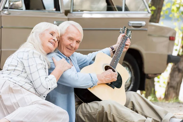 Senior Kobieta Siedziała Piasku Mężczyzną Grać Gitarze Przed Rocznika Samochodu — Zdjęcie stockowe