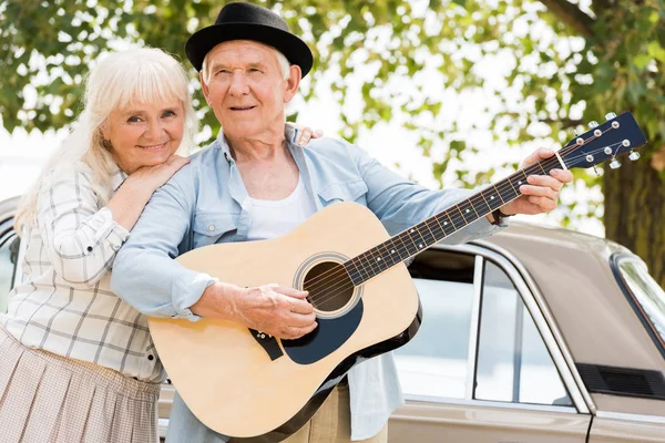 Senior Vrouw Knuffelen Echtgenoot Gitaarspelen Tegen Beige Auto — Stockfoto