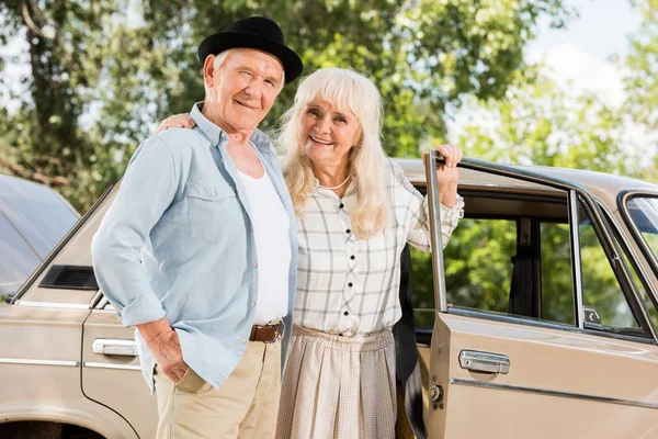 senior husband and wife standing near retro car and looking at camera