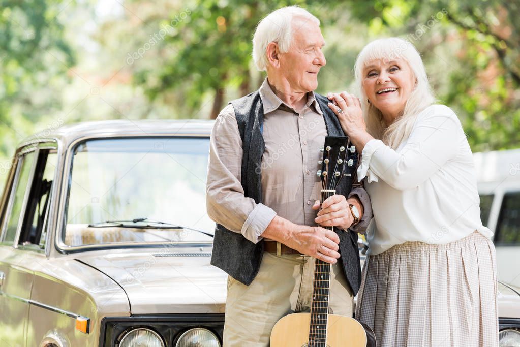 senior woman embracing man with guitar against beige vintage car 