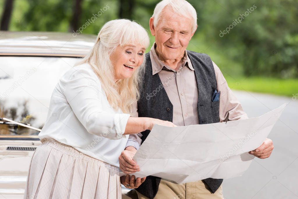 happy senior couple holding map and leaning on retro car