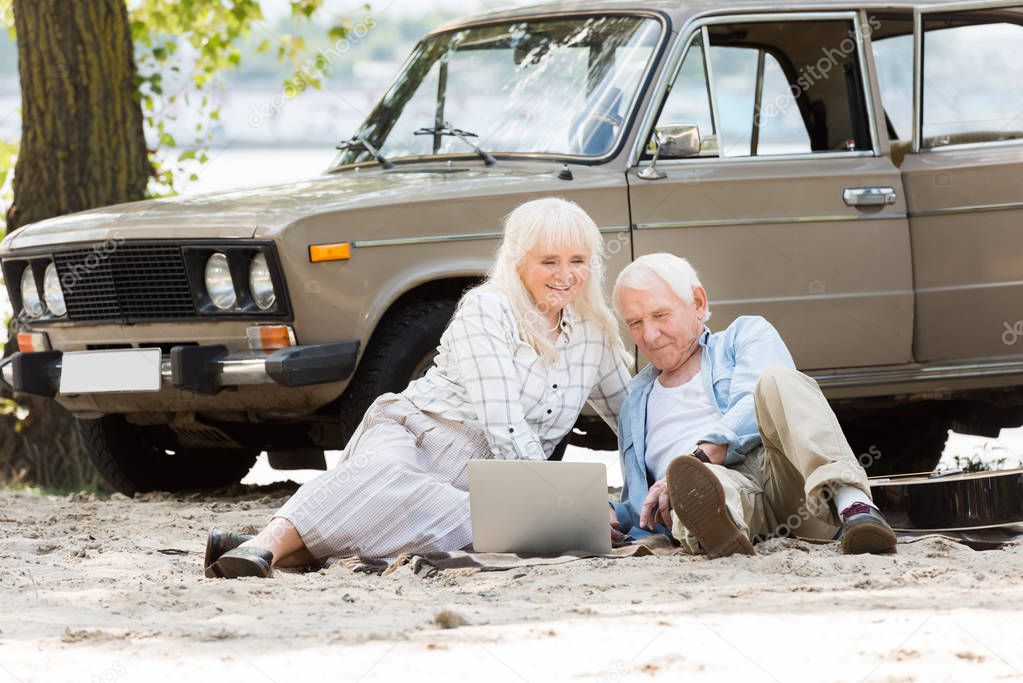 Happy senior couple sitting on sand and using laptop near beige vintage car